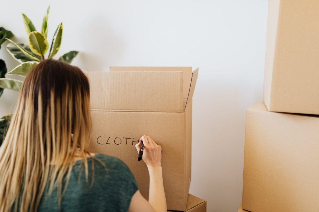 <woman labelling two boxes with the word “clothes”>