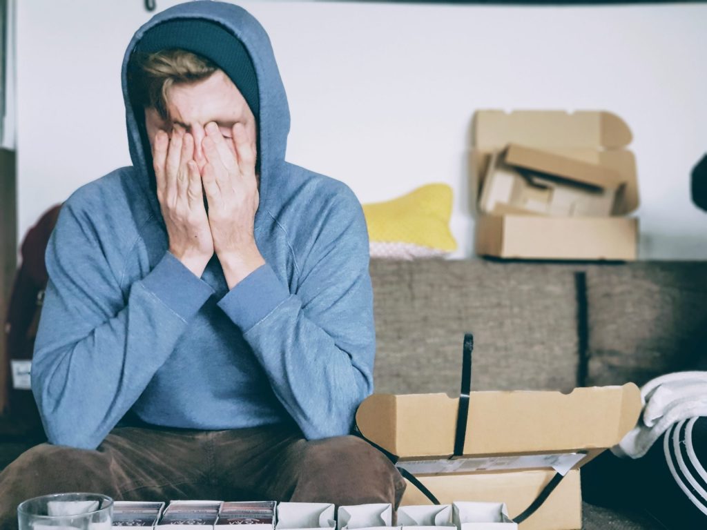 A young man covers his face in frustration as he sits on a sofa with boxes everywhere while he faces decluttering paralysis